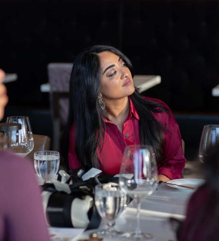 Woman in a red blouse sits at a dining table, looking to her left during a dinner with wine glasses nearby.